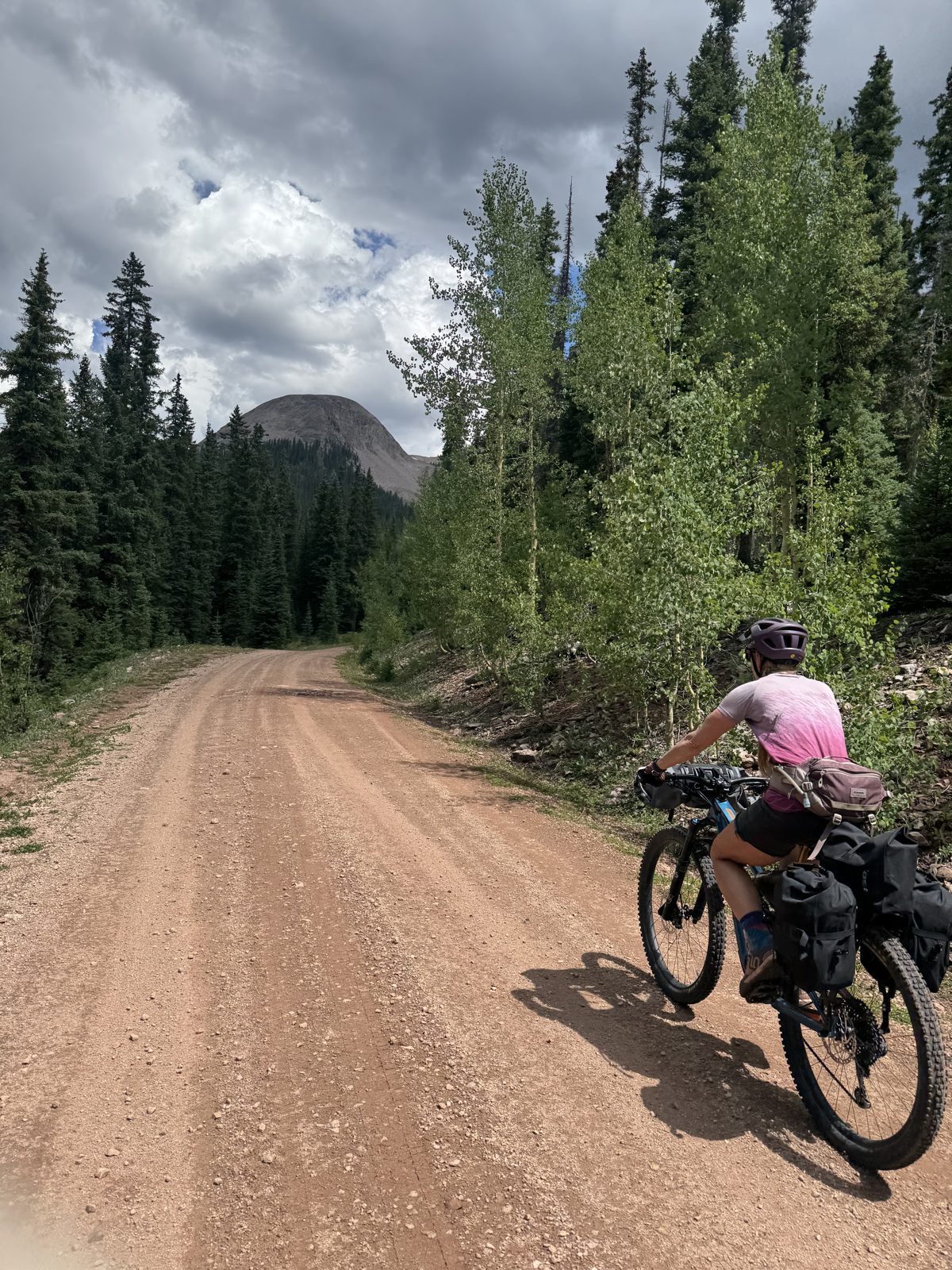 Cyclist riding a loaded mountain bike on a gravel road toward a bald mountain peak